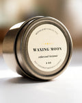 A round metal tin candle 'Waxing Moon' by Moonlight Collective, photographed on a clean white surface with a softly blurred warm-toned background.