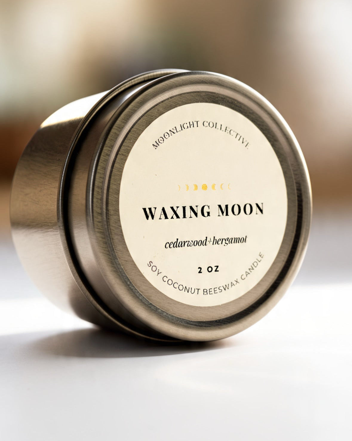 A round metal tin candle 'Waxing Moon' by Moonlight Collective, photographed on a clean white surface with a softly blurred warm-toned background.
